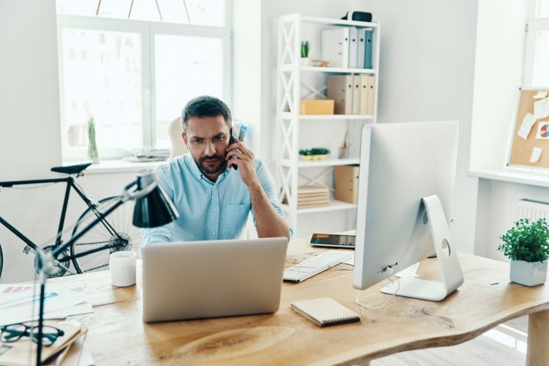 Handsome middle age remote worker in smart casual wear using laptop and talking on the phone while sitting in his home office